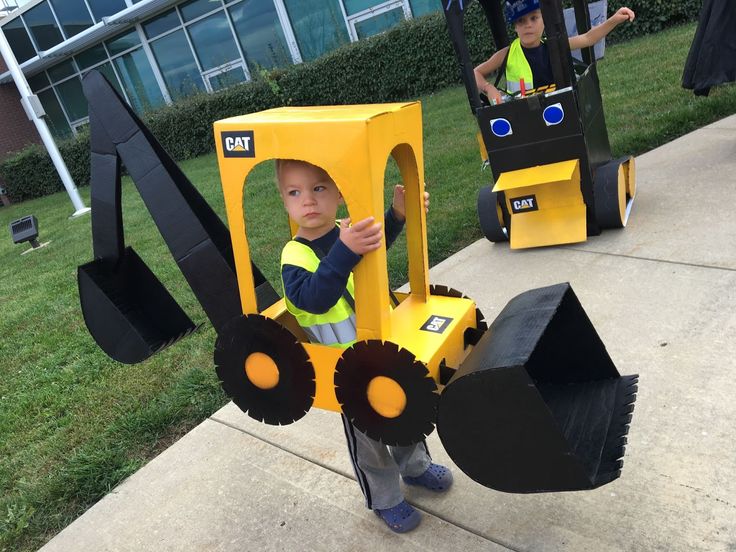 two children in construction costumes standing on the sidewalk with large equipment and one child wearing a construction vest