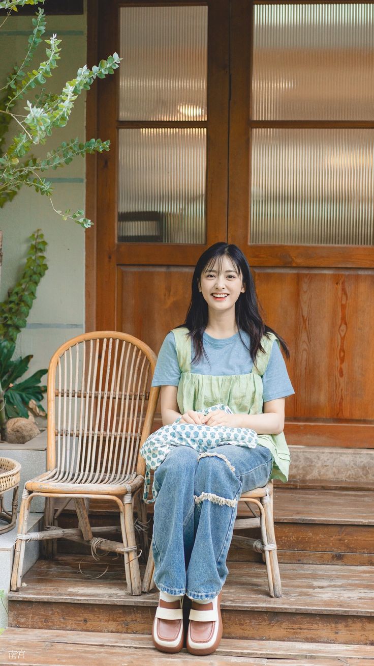 a woman sitting on a porch next to a chair and potted plant in front of a wooden door