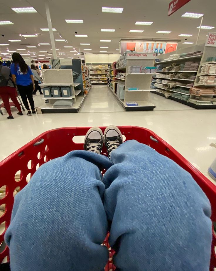 someone is laying down in a red shopping basket at the grocery store with their feet propped up