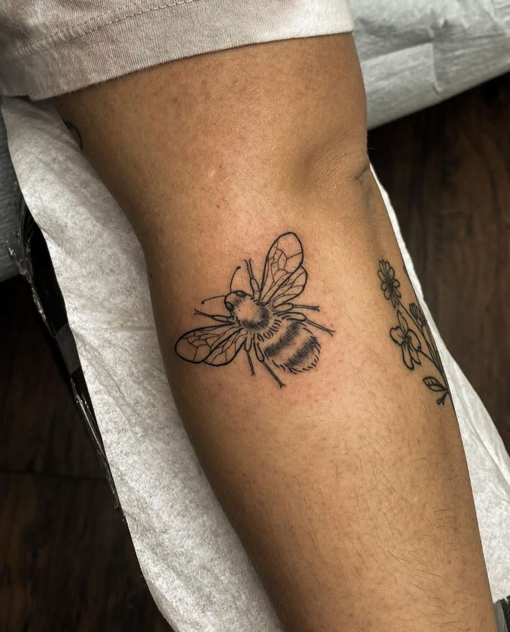 a small bee tattoo on the right arm and left leg, with flowers around it