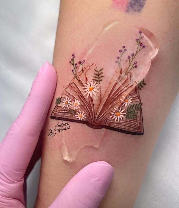 a person with tattoos on their arm holding an open book and flowers in the pages