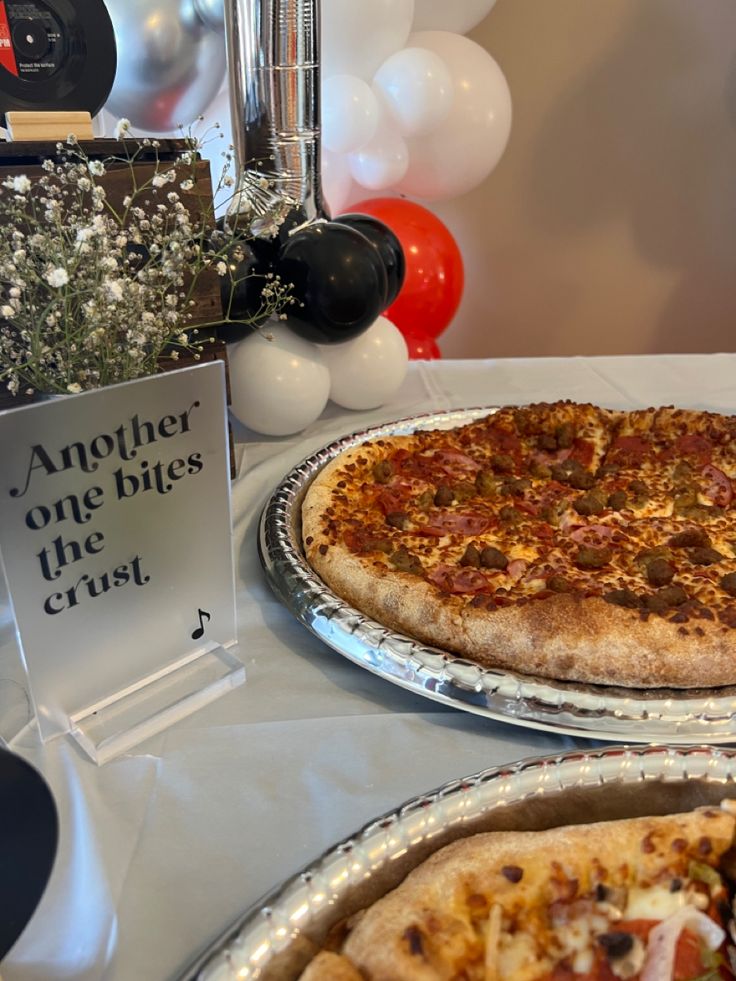 two pizzas on pans sitting on a table with balloons and decorations in the background