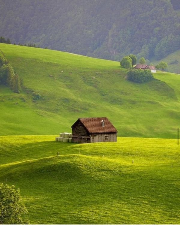 an old barn sits in the middle of a green field with rolling hills behind it