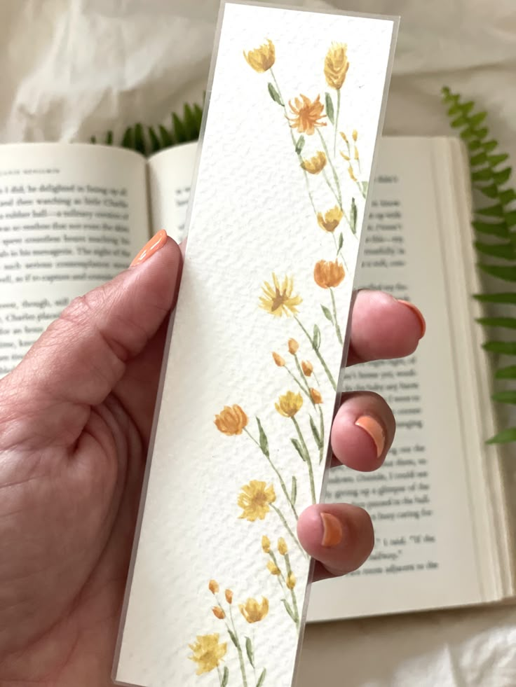 a hand holding an open book with watercolor flowers on it and a fern in the background