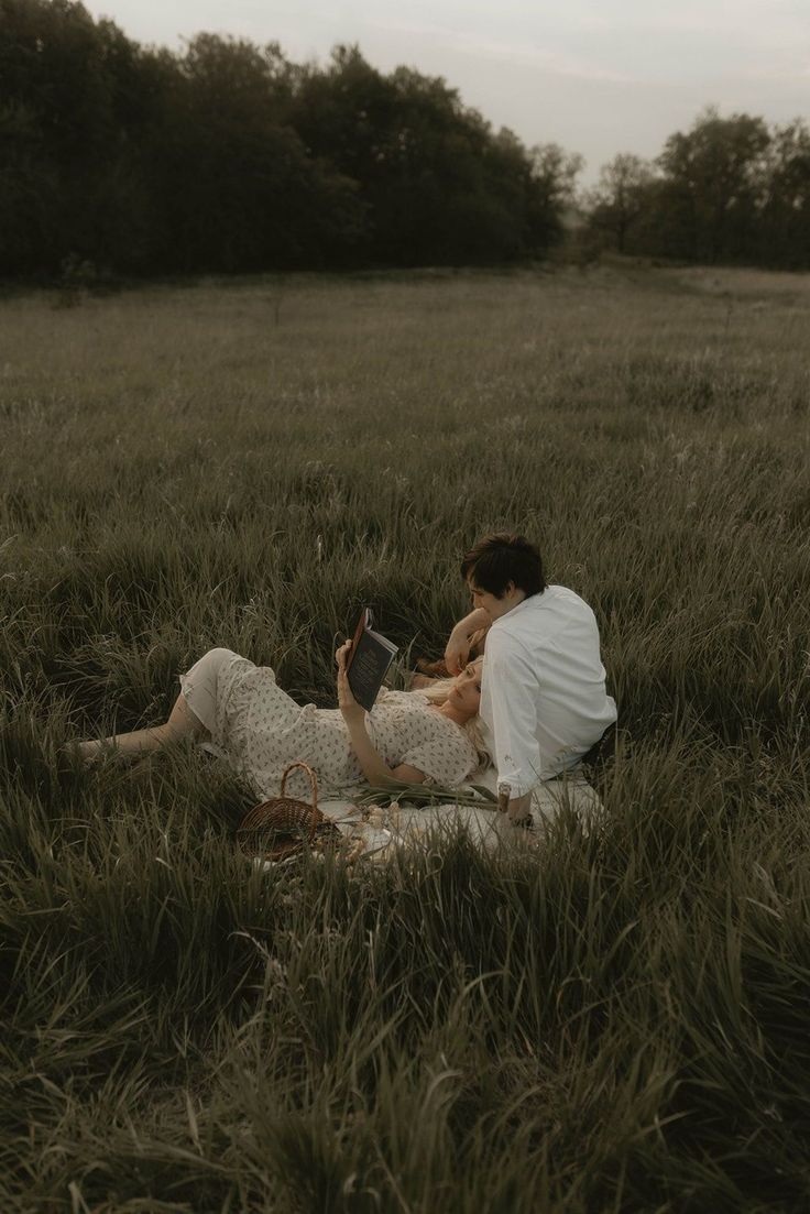 a man and woman sitting on the ground in a grassy field with trees behind them