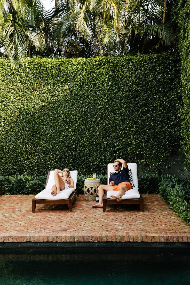 two people lounging on lounge chairs next to a swimming pool in front of a green hedge