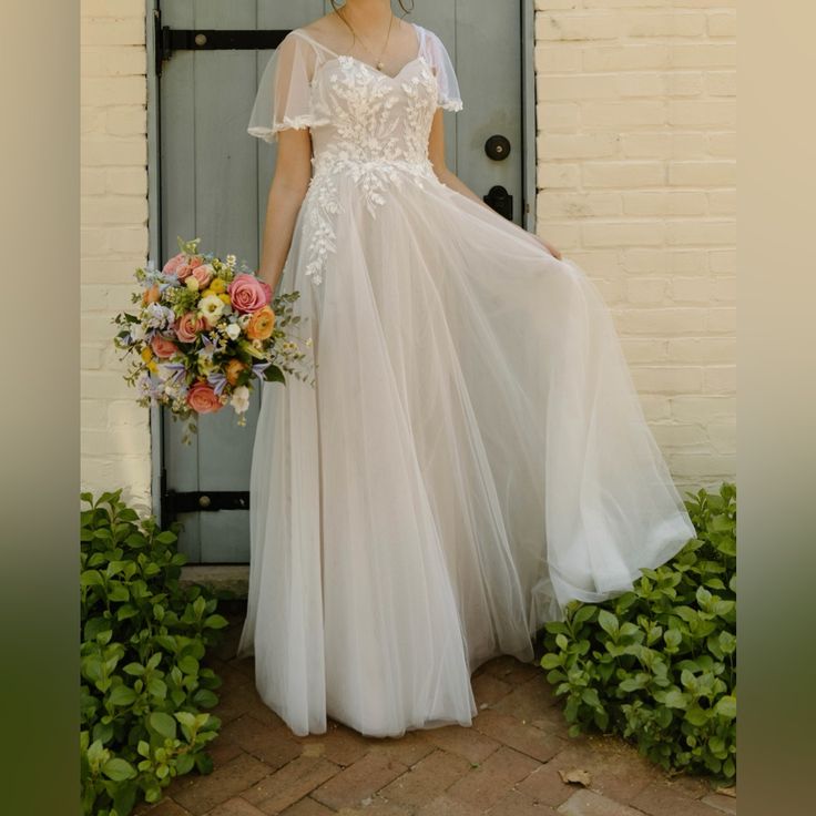 a woman standing in front of a door wearing a white dress and holding a bouquet