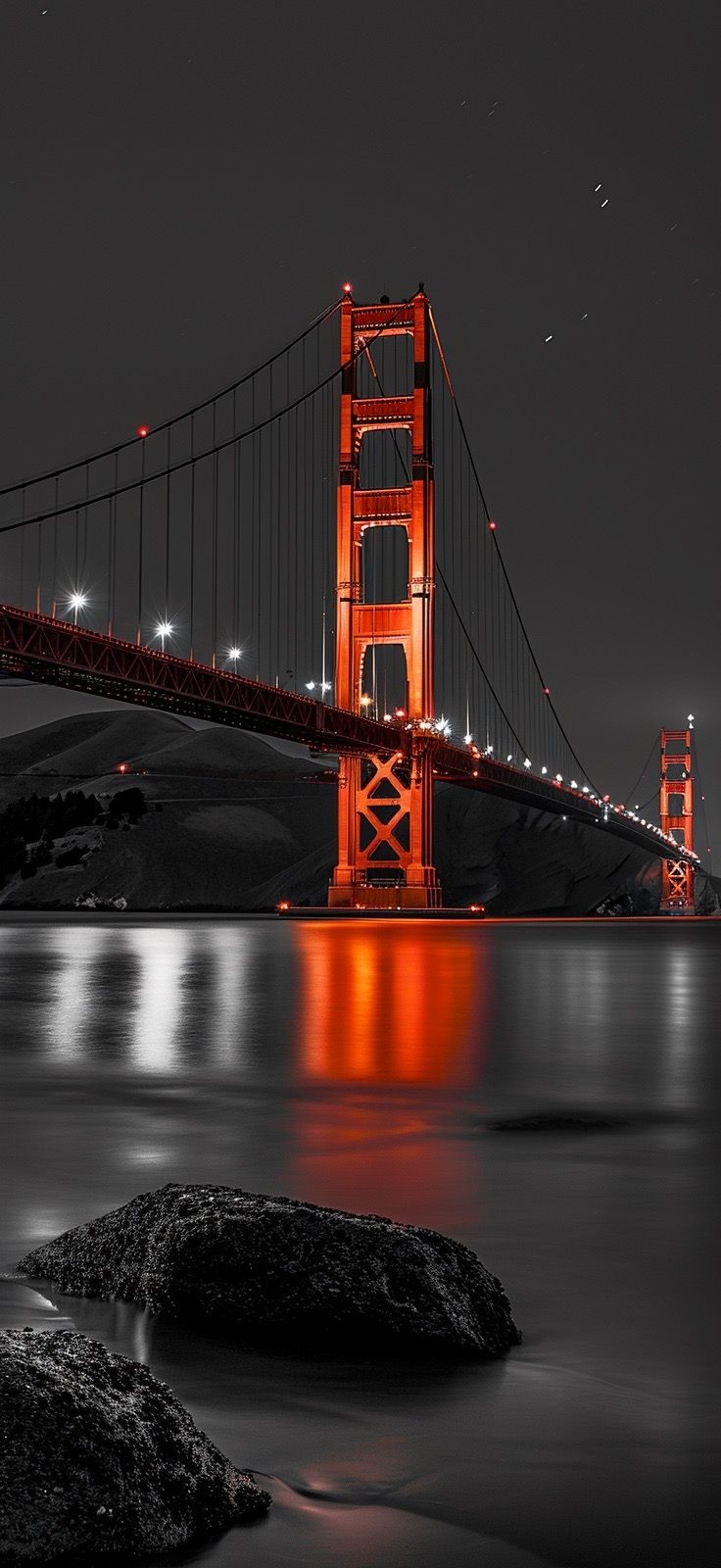 the golden gate bridge lit up at night with lights reflecting off water and rocks in foreground