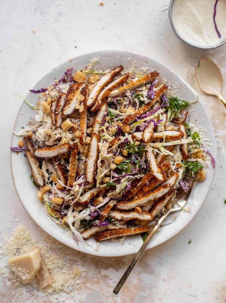 a white plate topped with chicken and coleslaw next to a bowl of dressing