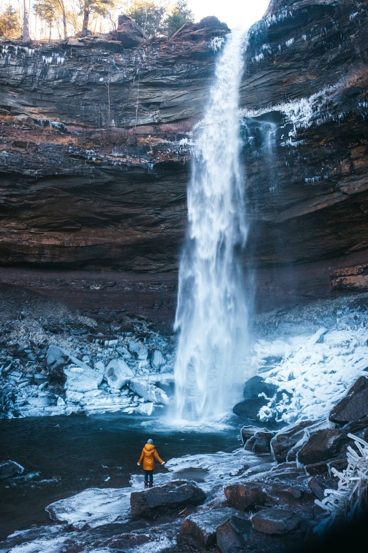 a person standing in front of a waterfall with snow on the ground and rocks below