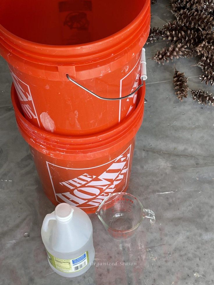 two orange buckets sitting next to each other on top of a cement floor with pine cones in the background