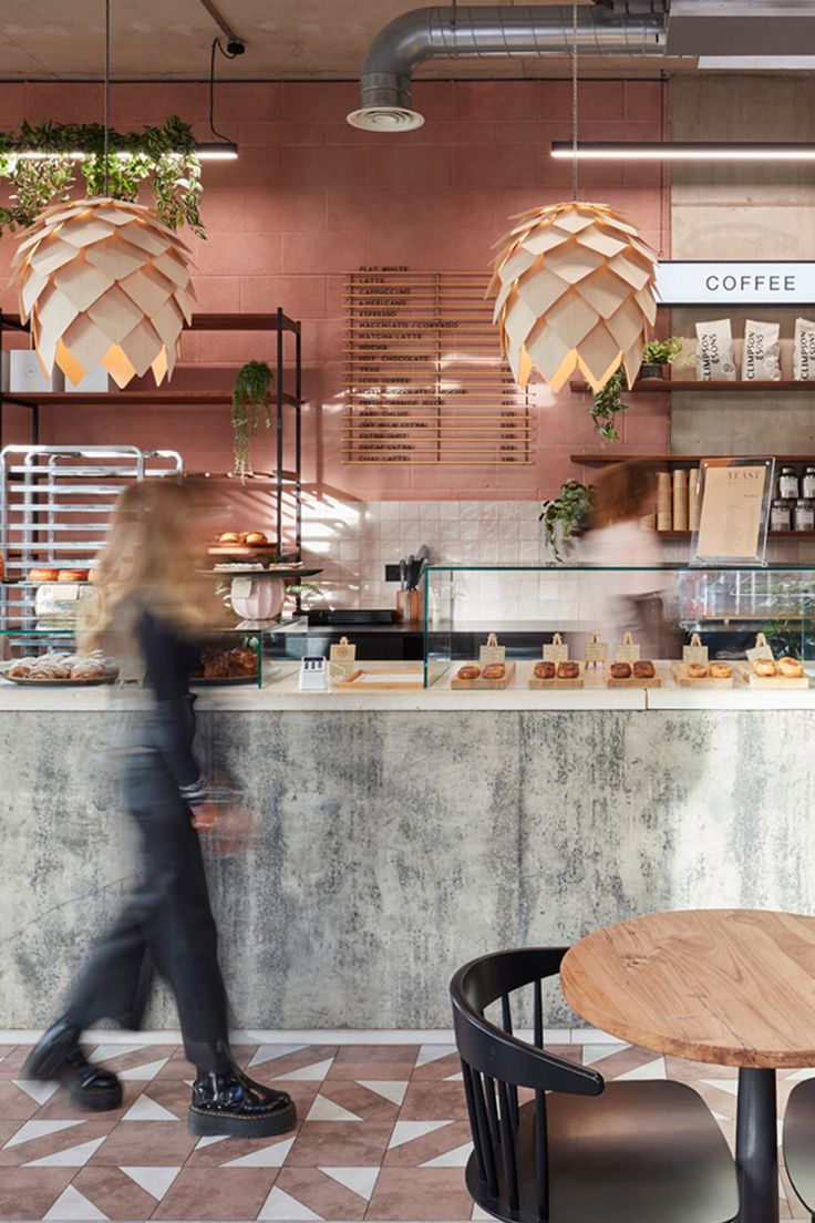 a woman walking past a counter in a coffee shop with lots of plants hanging from the ceiling