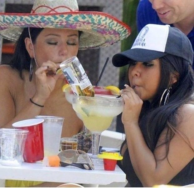 two women sitting at a table with drinks and hats on, one drinking from a straw