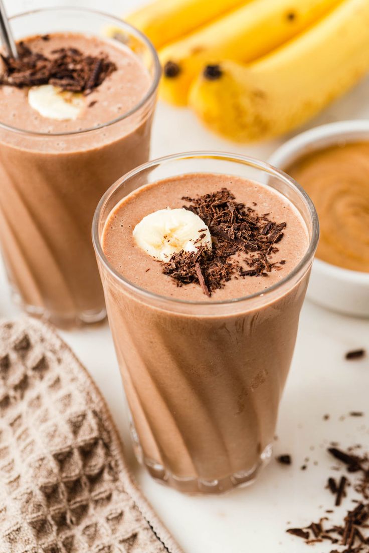 two glasses filled with chocolate smoothie next to banana's and other ingredients on a white surface