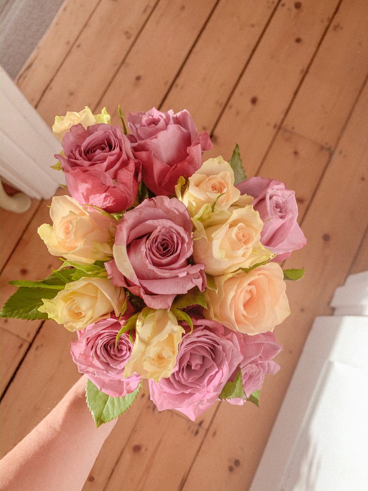 a bouquet of pink and yellow roses in someone's hand on a wooden floor