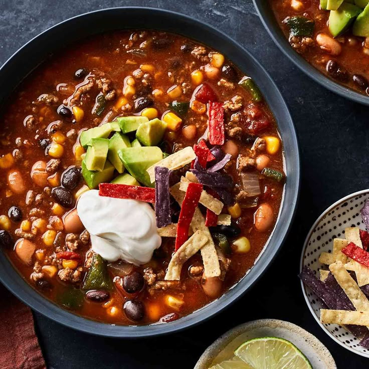 two bowls of chili and beans with tortilla chips, avocado wedges and sour cream