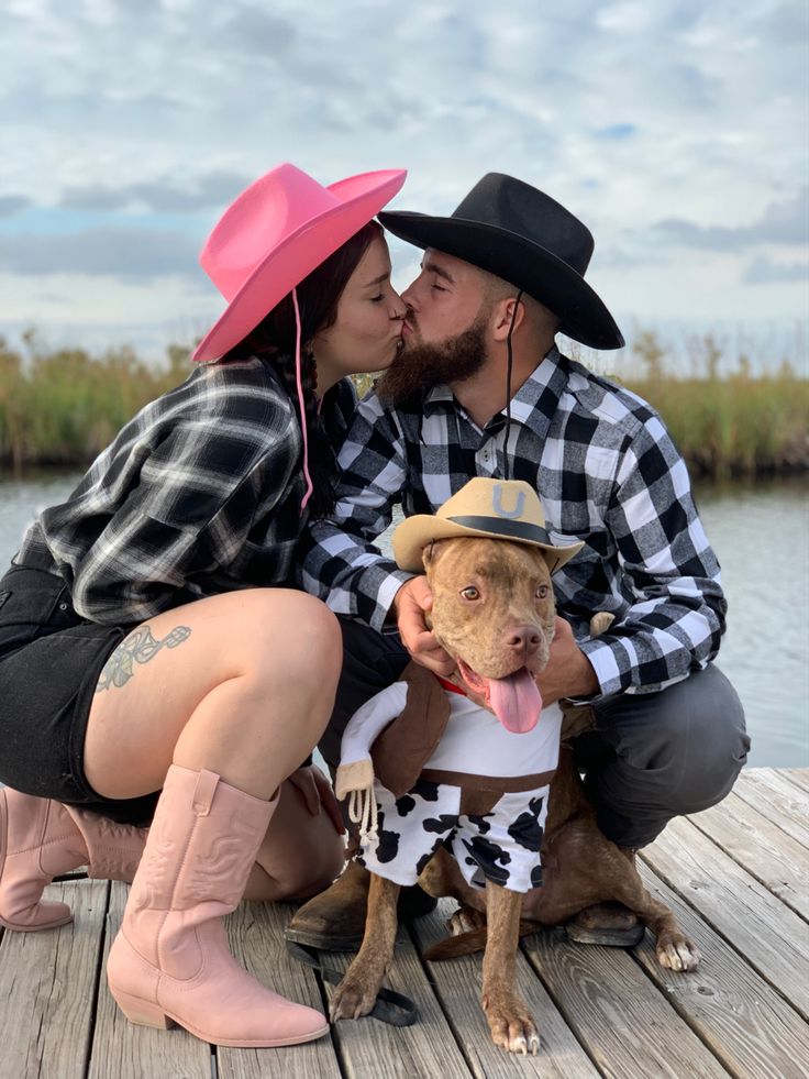 a man and woman kissing while sitting on a dock with a dog wearing a cowgirl outfit