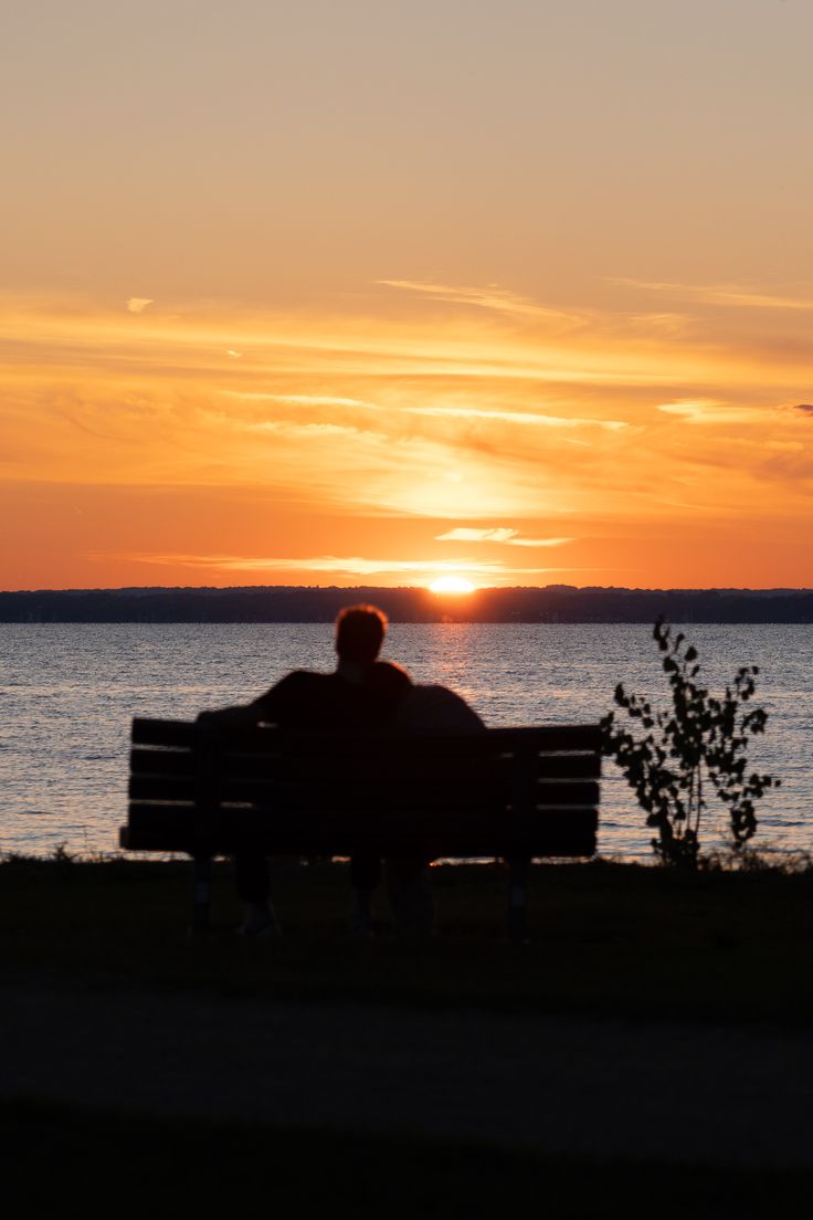 a man sitting on top of a bench next to the ocean at sunset or dawn