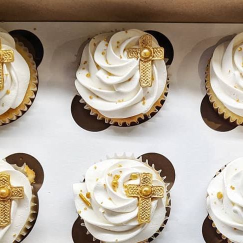 twelve cupcakes with white frosting and gold decorations in a box, each decorated with a cross