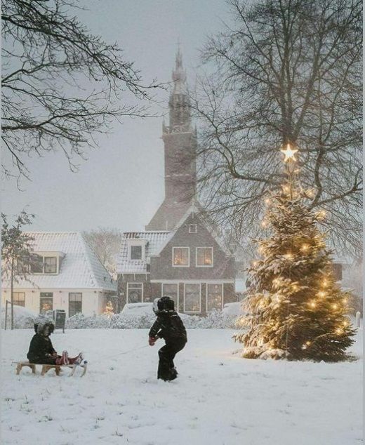 two people walking in the snow near a christmas tree and a church with lights on it