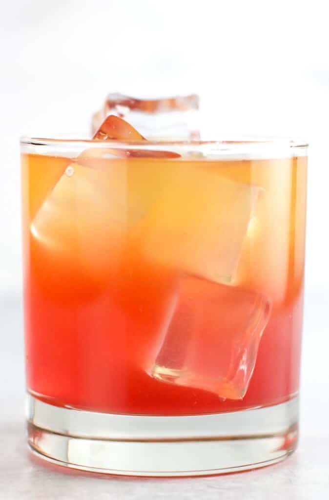 an orange and red drink in a glass with ice cubes on the rim, sitting on a white surface