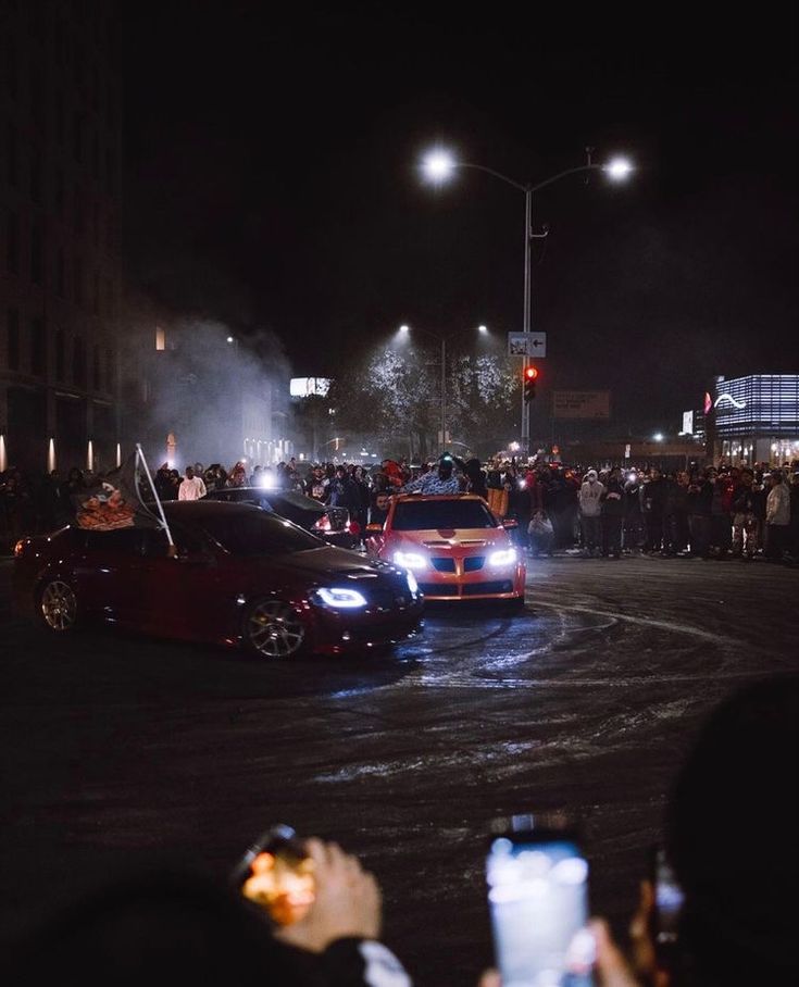 two cars driving down a street at night with people standing on the side of the road