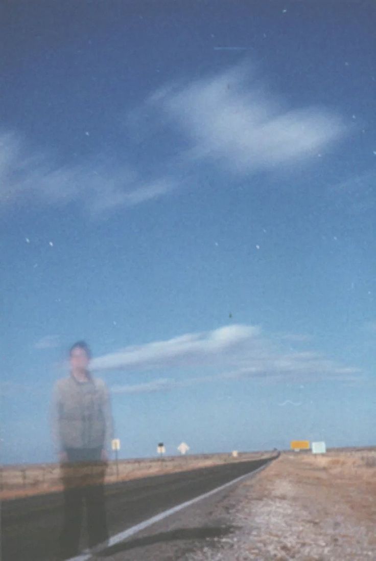 a man standing on the side of a road in front of a sky filled with clouds