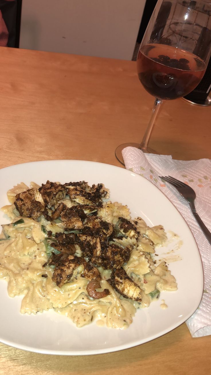 a white plate topped with pasta and meat next to a glass of wine on a wooden table