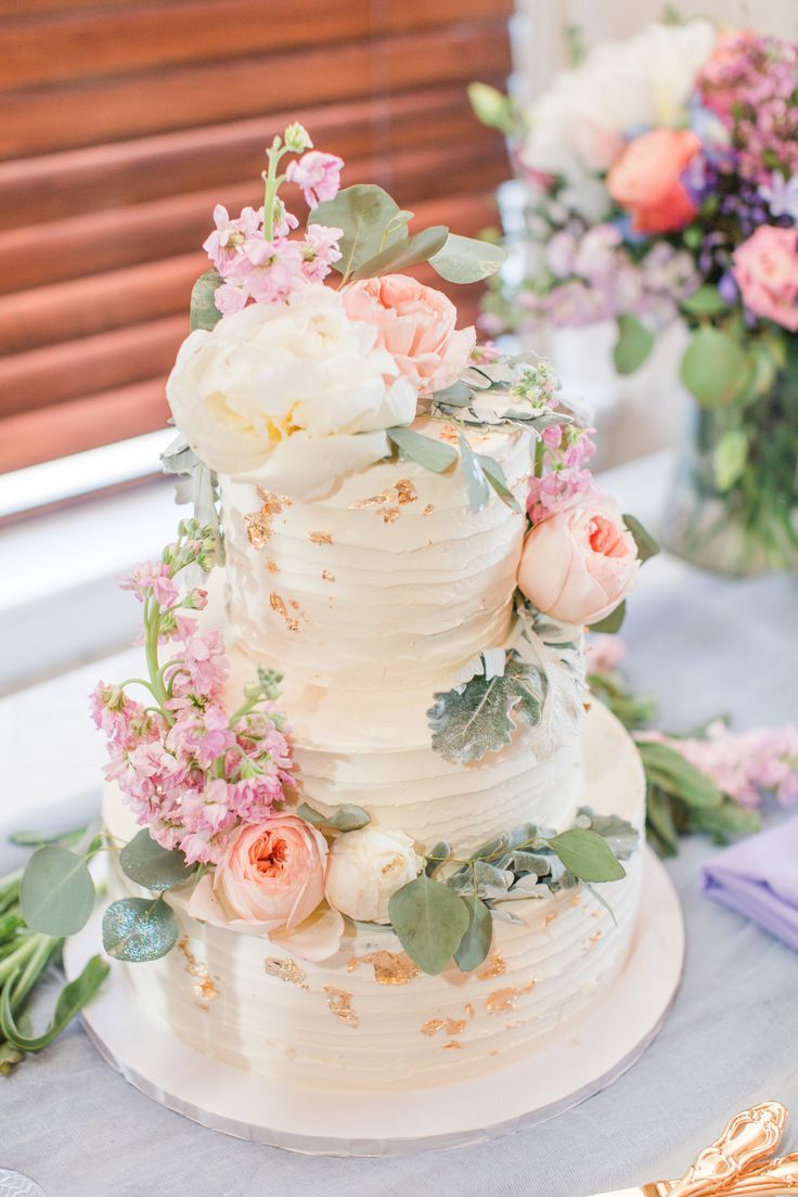 Spring wedding cake at reception. White 3 tiered buttercream with soft pastel colored flowers on the frosting. Bridgerton Pastel Wedding Theme, Aesthetic Wedding Cake Korean, White Wedding Cake Pink Flowers, Spring Pastel Wedding Cake, Pastel Wedding Flowers Table, Peonies Wedding Cake, Spring Themed Wedding Cake, Pastel And Greenery Wedding, Peony Themed Wedding