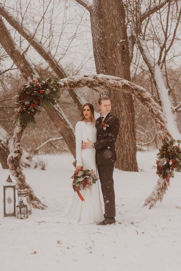 a bride and groom standing in the snow under an arch made of branches with flowers