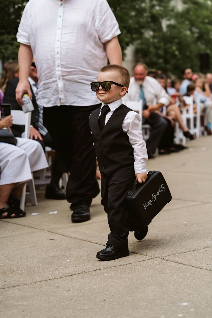 a little boy in a suit and sunglasses walking down the street while holding a briefcase