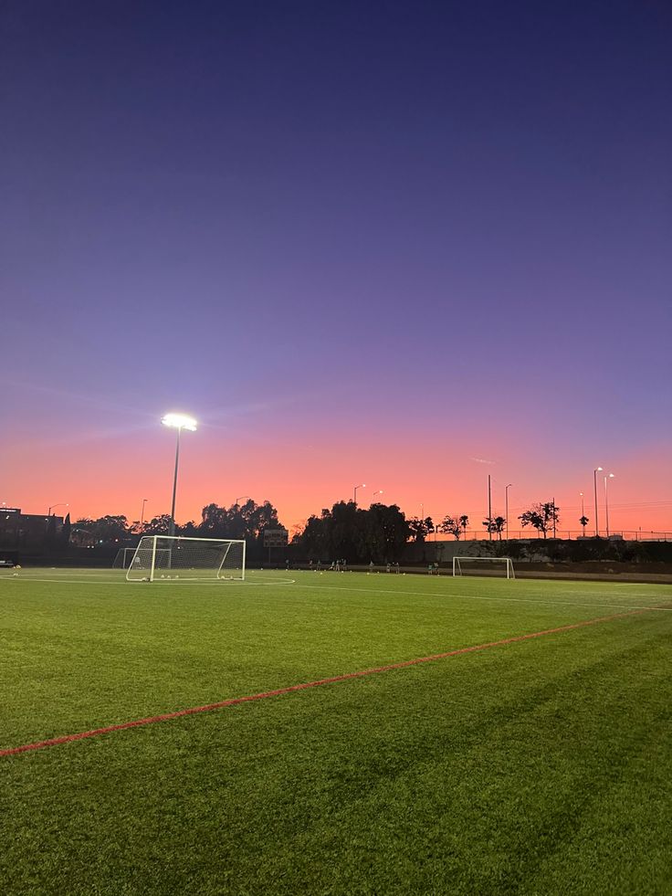 an empty soccer field at sunset with the sun going down in the distance and lights on