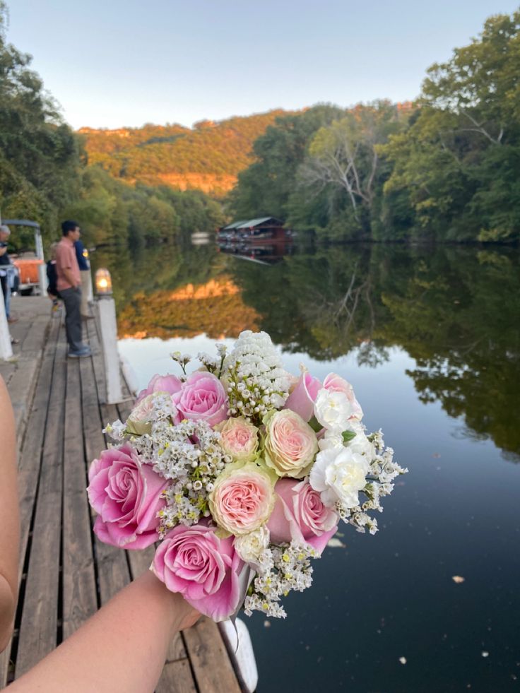 a person holding a bouquet of pink and white flowers on a dock over water with people in the background