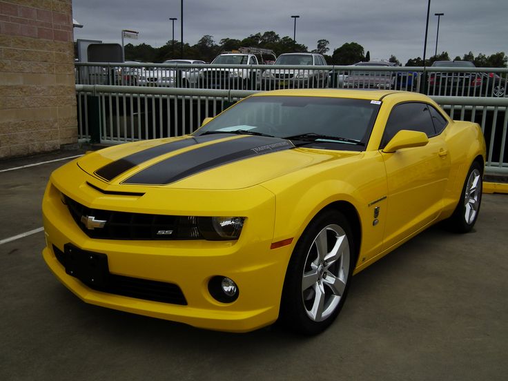 a yellow sports car with black stripes parked in a parking lot next to a fence