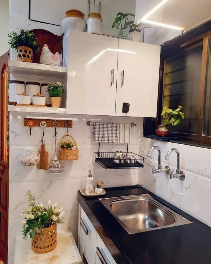 a small kitchen with white cabinets and black counter tops, plants on the shelf above the sink