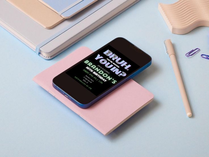 a phone sitting on top of a pink notebook next to a pen and laptop computer