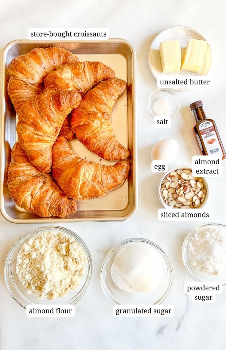 the ingredients to make croissants laid out on a white counter top, including butter, eggs, almonds, flour, and sugar