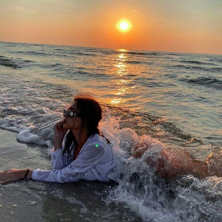 a woman laying on the beach with her feet in the water and sun setting behind her