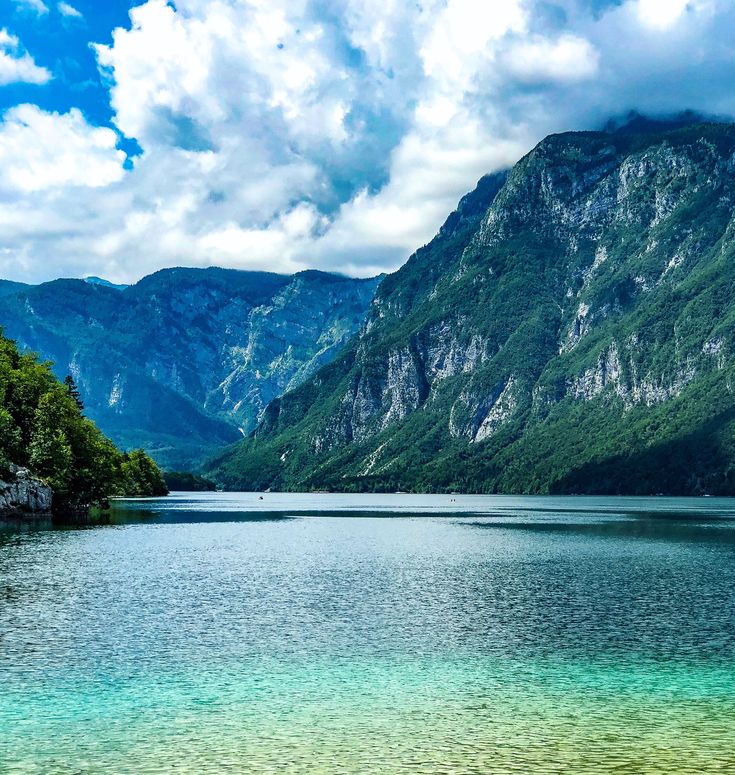 a lake surrounded by mountains under a blue sky with clouds in the background and green trees on both sides