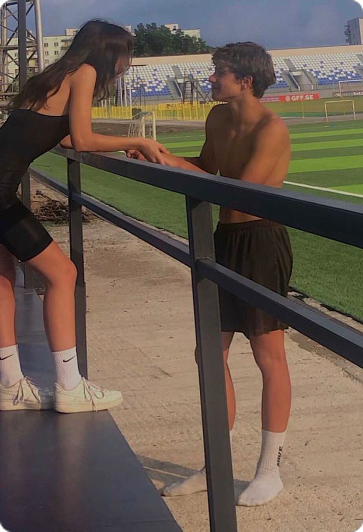 a man and woman leaning against a rail at a baseball field with the stadium in the background