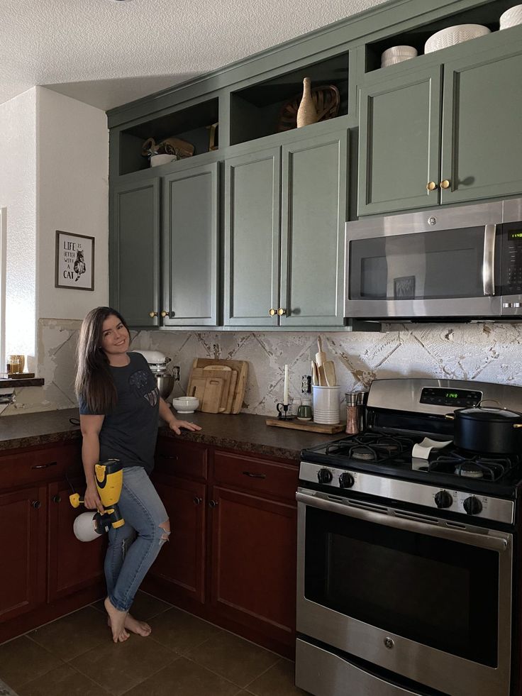 a woman standing in a kitchen with green cabinets and silver stove top oven, holding a yellow mitt