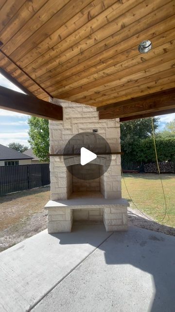 an outdoor fireplace in the middle of a patio with a wooden roof and brick wall