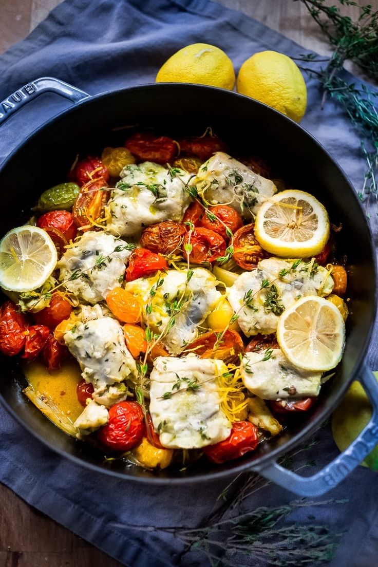 a pan filled with chicken and tomatoes on top of a blue cloth next to lemons