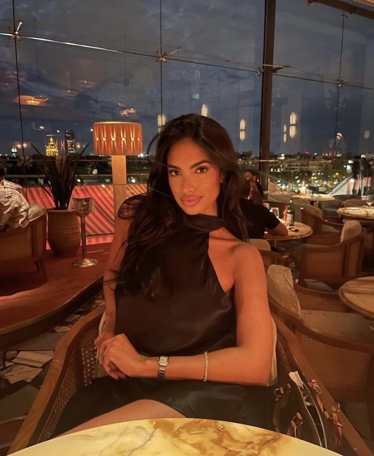 a beautiful woman sitting at a table in front of a window with the city lights behind her