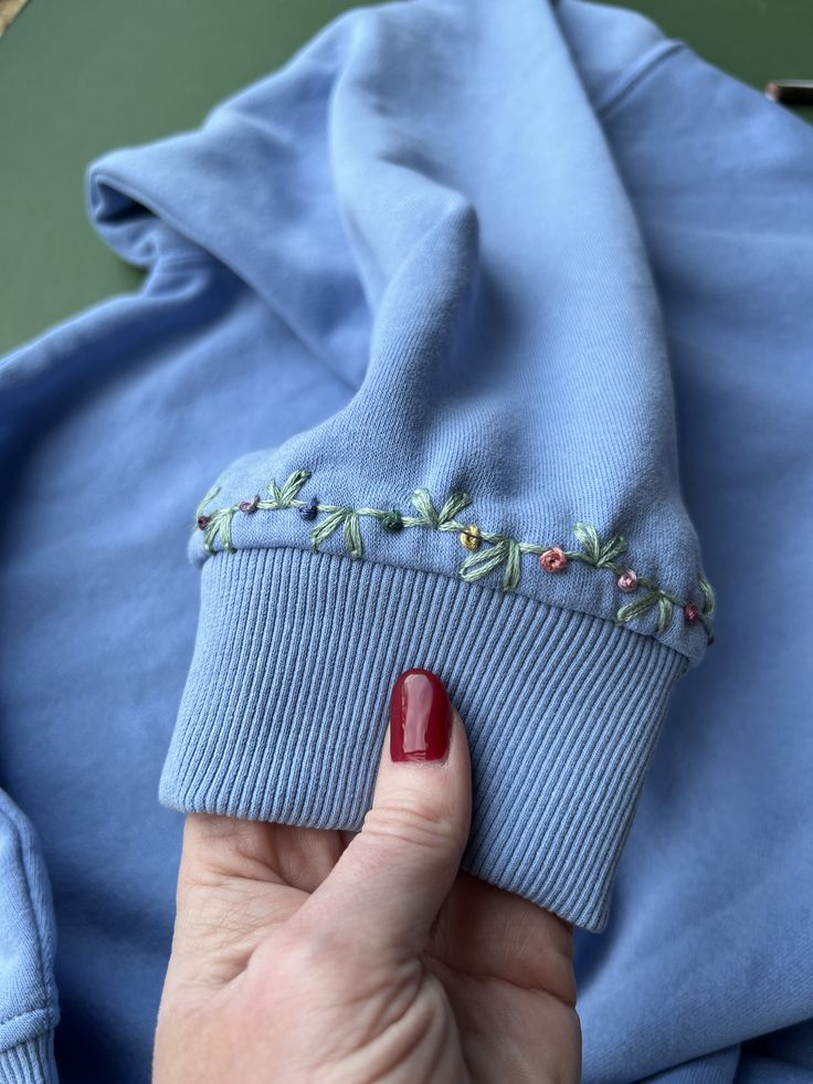 a woman's hand holding onto a blue sweater with embroidered flowers on the sleeves