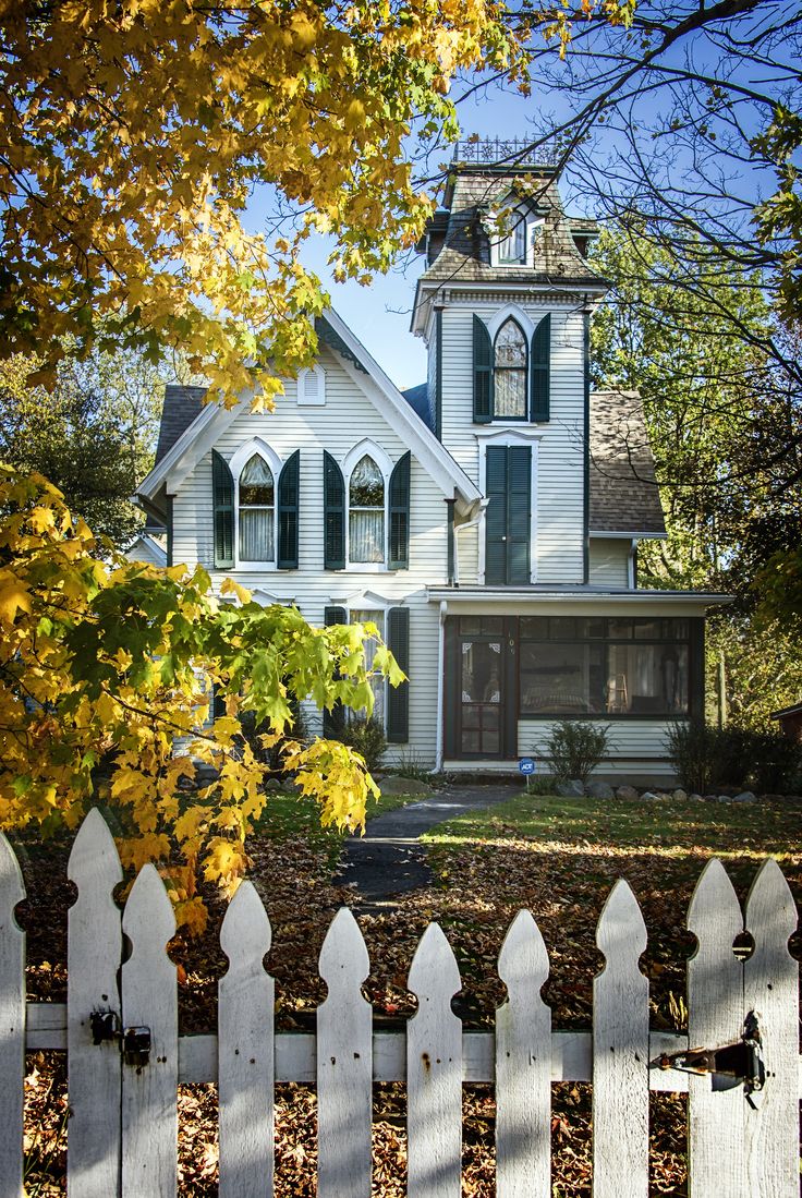 a white picket fence in front of a house with autumn leaves on the ground and trees around it