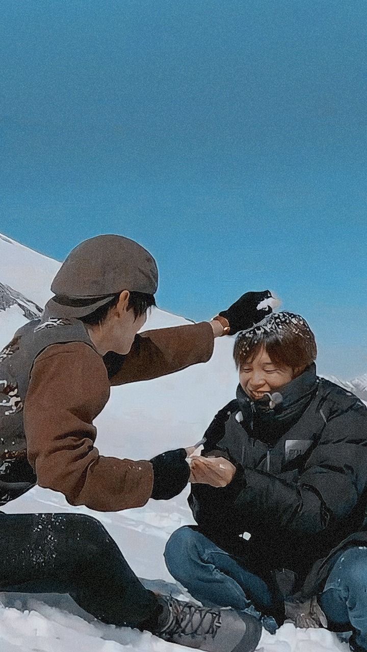 two people sitting in the snow with one touching another's ear while they both look at their cell phones