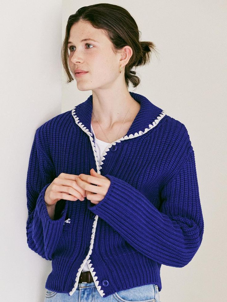 a woman standing in front of a white wall wearing a blue cardigan sweater and jeans