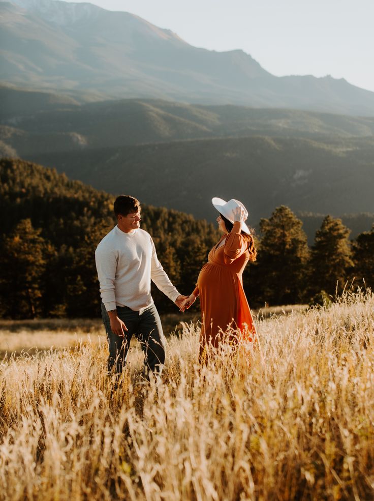 a man and woman holding hands while walking through tall grass in front of mountain range