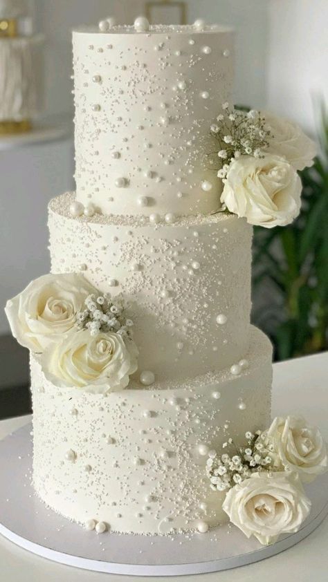 a three tiered wedding cake with white roses on top and pearls around the edges